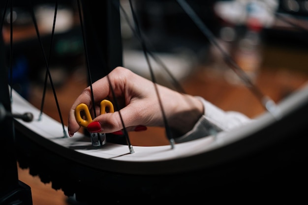 Foto colpo ritagliato del primo piano delle mani irriconoscibili del riparatore di ciclismo femminile che controllano il raggio della ruota della bicicletta con la chiave del raggio che lavora nell'officina di riparazione con interni scuri