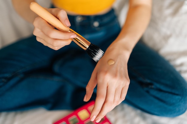 Closeup cropped shot of unrecognizable female put makeup foundation on hand and going to use brush to apply makeup to face sitting on bed in bedroom