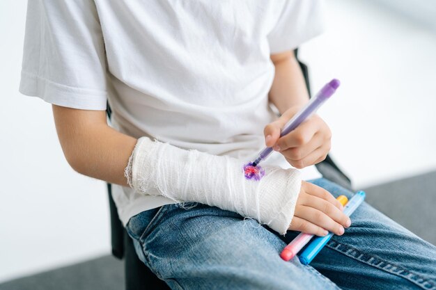 Closeup cropped shot of unrecognizable little girl drawing cute\
image on cast of broken arm wrapped in white plaster bandage on\
white isolated background