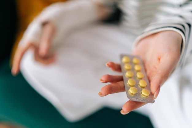 Closeup cropped shot of unrecognizable suffering from pain young woman with broken right hand wrapped in gypsum bandage holding blister packs of pills