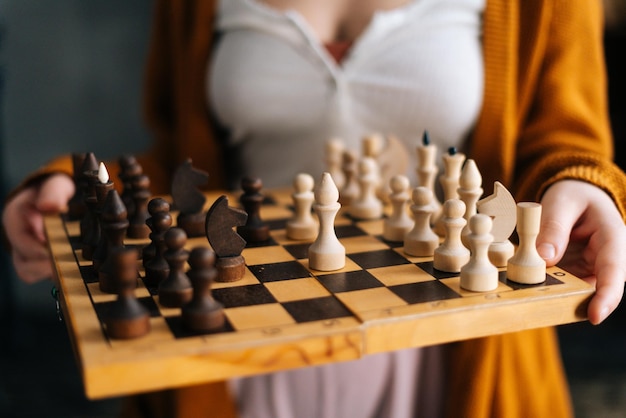 Closeup cropped shot of unrecognizable young woman holding in\
hands chess board standing on vintage wooden floor in dark\
room