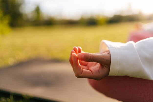 Closeup cropped shot of unrecognizable young woman meditating sitting in lotus position on yoga mat outside in park on background of sunray and green grass