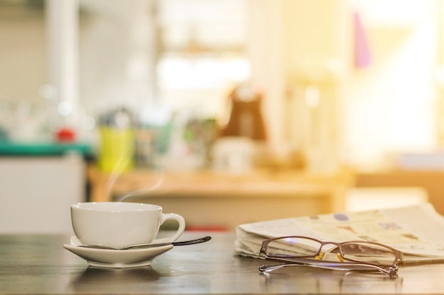 Closeup cup of hot cappuccino coffee with newspaper and spectacles on the wood table.