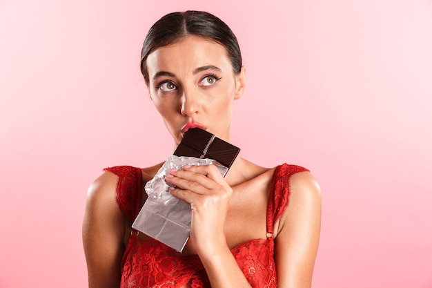 Photo closeup cute thinking woman wearing red sexual lace lingerie eating chocolate bar closed isolated over pink