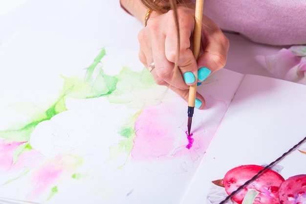 Closeup darkhaired beautiful artist with bright nails draws a thin wooden brush and watercolor in the album for drawing on white paper pink peonies