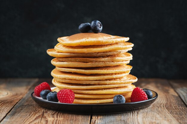 Closeup delicious pancakes with fresh blueberries raspberry and maple syrup or honey on a dark background Sweet maple syrup flows from a stack of pancake