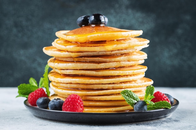 Closeup delicious pancakes with fresh blueberries raspberry and maple syrup or honey on a dark background Sweet maple syrup flows from a stack of pancake