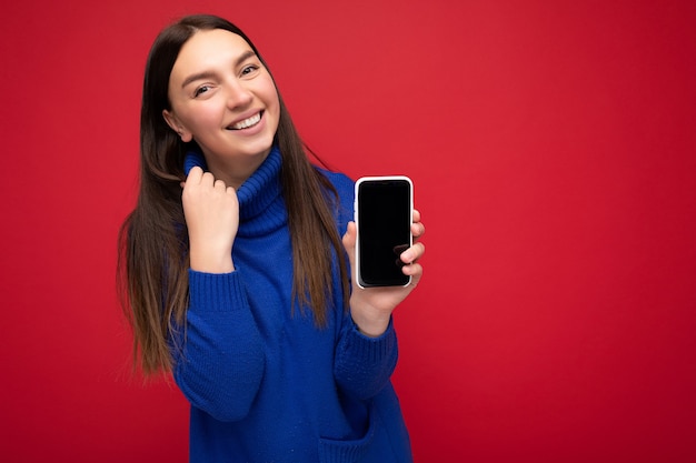 Closeup of delightful beautiful happy young brunette woman wearing casual blue sweater isolated over red background with empty space holding in hand mobile phone and showing smartphone with empty scre