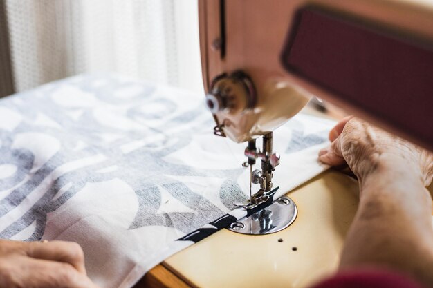 Closeup detail of wrinkled hands of old woman sewing fabric\
patterns on her sewing machine at home