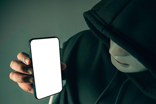 CloseUp Een anonieme hacker met een masker en een zwarte hoodie pronkt met een smartphone met een leeg wit scherm Hacking en Malware Concepts