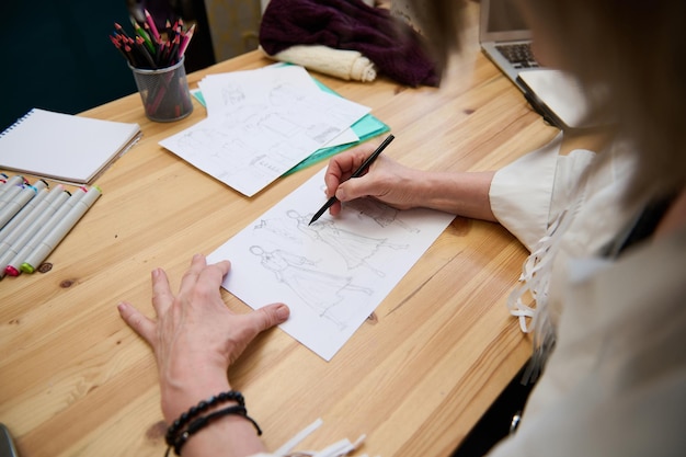高齢者に触発されたファッションデザイナーのクローズアップは、木製のパーソナルデスクで鉛筆と水彩画のマーカーを使用してラインを作成するデザインスタジオの女性の仕立て屋で新しい服のコレクションのスケッチを描く