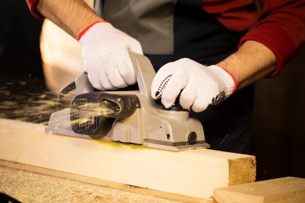 Closeup of electric plane throws sawdust while woodworker making wooden detail on workbench at cottage workshop