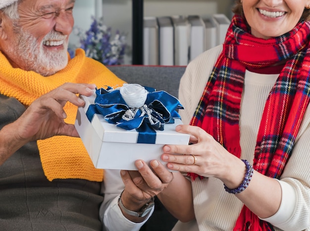 Closeup exchanging christmas present or gift in hands of happy\
caucasian senior couple during the day at home. christmas and new\
year celebration activity.