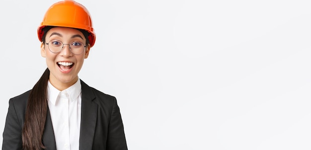 Closeup of excited upbeat asian female chief construction engineer architect in suit and safety helmet smiling amazed looking with hope and satisfaction at good work done white background