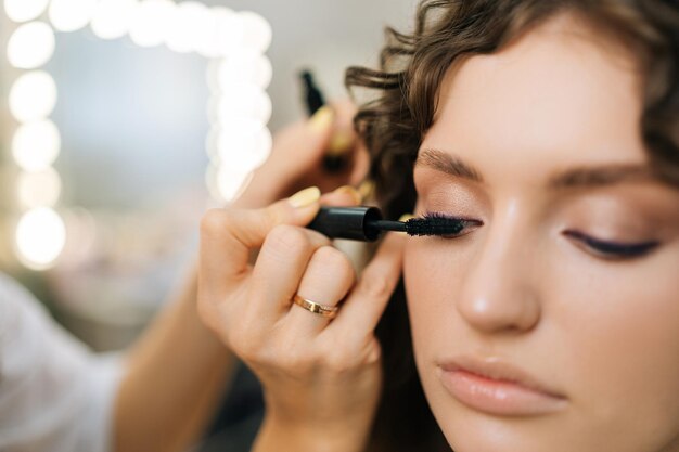 Близкий взгляд на привлекательную молодую женщину, наносящую маскару на ресницы от макияжа в салоне красоты на размытом фоне зеркала
