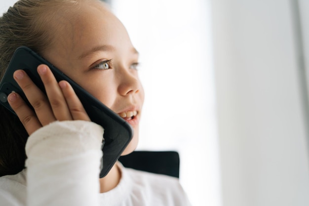 Closeup face of cheerful blonde little girl with broken arm wrapped in plaster bandage talking smartphone smiling looking away on white background