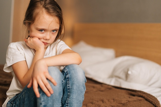 Closeup face of frowning beautiful little girl with wet eyes from tears sits on bed in bedroom and looking down