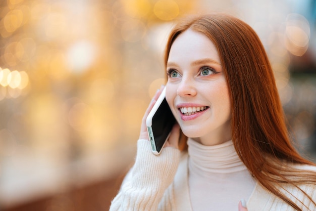 Closeup face of happy attractive redhead young woman talking on mobile phone standing in shopping mall with bright interior during holiday sales looking away blurred background bokeh lights
