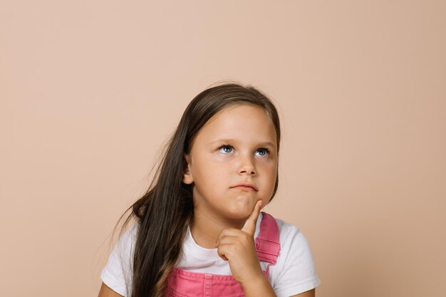 Closeup face kid with shining bright eyes looking upward point finger on chin area of face with pensive look wearing bright pink jumpsuit and white tshirt on beige background