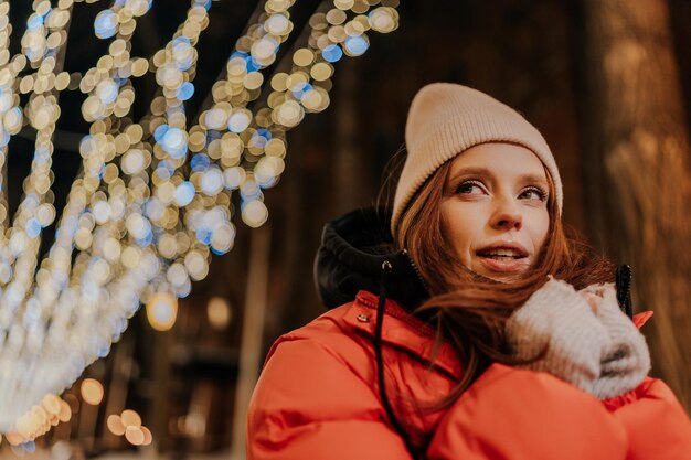 Closeup face of pretty frozen young woman in hat and winter jacket warming hands in cold winter