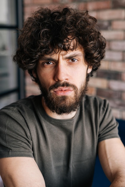 Closeup face of serious handsome young man looking at camera sitting on chair in cozy living room with brick wall