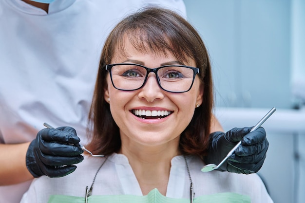 Closeup face of smiling mature woman with teeth in dentistry with hands with dentist examination tools Treatment prosthetics implantation whitening hygiene dental health care concept