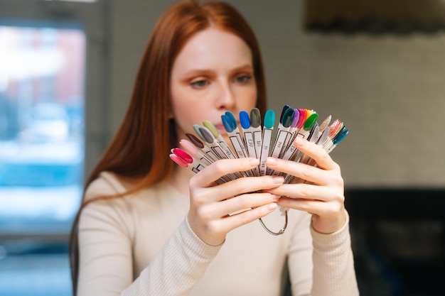 Closeup face of thoughtful pretty young woman client choosing colour from colourful nails tips in manicure salon Serious attarctive redhead female choosing perfect nail varnish color