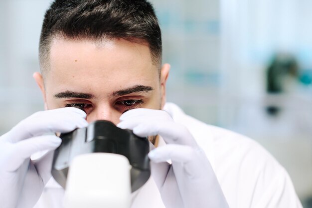 顕微鏡で見ている手袋の若い深刻な男性科学者の顔のクローズアップ