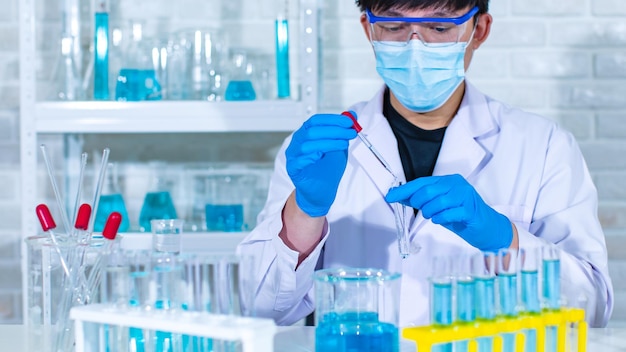 Снимок лица крупным планом азиатского молодого профессионального ученого-мужчины в белом лабораторном халате в защитных очках с помощью объектива микроскопа, смотрящего на образец микробиологии на стеклянной пластине.