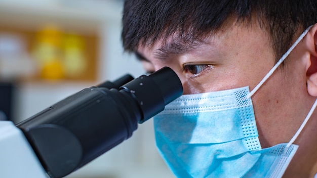 Closeup facial shot of Asian young professional male scientist in white lab coat wearing safety protection goggles glasses using microscope lens looking zooming at microbiology sample on glass plate.