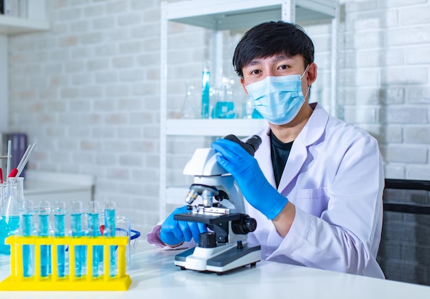 Closeup facial shot of asian young professional male scientist in white lab coat wearing safety protection goggles glasses using microscope lens looking zooming at microbiology sample on glass plate