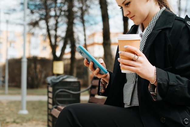 Closeup of fashionable young woman chatting on internet on smartphone while sitting on park bench Side view of pretty lady holding mobile phone and cup of coffee outdoors Selective focus on phone