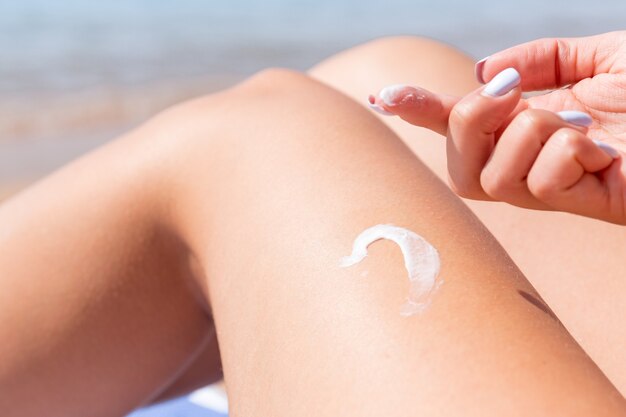 Крупным планом на женской руке, применяя крем для загара на ее загорелой ноге на пляже.