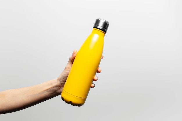 灰色の背景に黄色の金属製の再利用可能な熱水のボトルを持っている女性の手のクローズ アップ