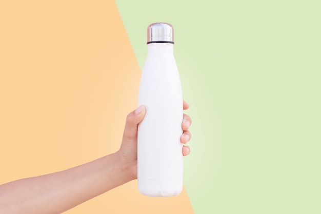 Closeup of female hand holding white reusable steel thermo water bottle isolated on two background of orange and green colors.