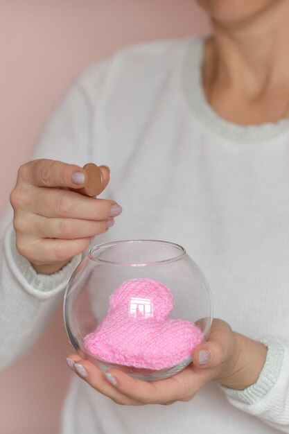 closeup female hands hold a transparent glass jar with a knitted heart inside and a coin