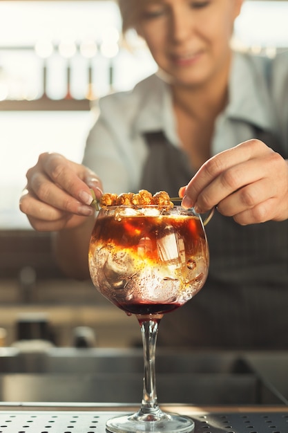 Closeup of female hands making alcohol cocktail in glass with ice cubes in cafe. Small business and professional barman concept