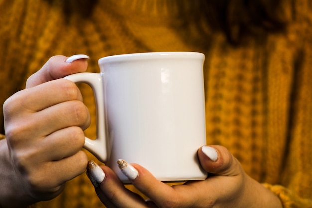 お茶のカップを保持しているエールセーターで飲み物の美しい女性のマグカップと女性の手のクローズアップ