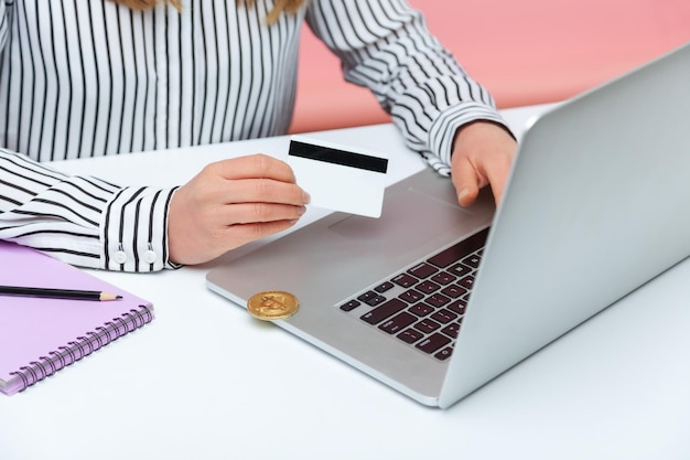 Closeup female holding credit card typing on laptop bitcoin crypto coin lying on pc corner withdrawal of electronic money to the card banking Indoor studio shot isolated on pink background