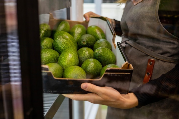 Closeup female kitchen staff hands in apron choosing fresh\
green avocado from cardboard box