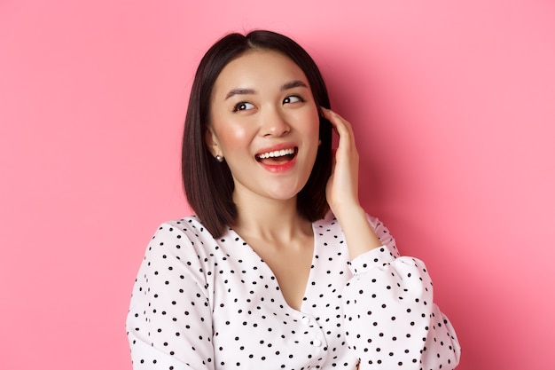 Closeup of flirty asian woman sighing looking dreamy left and smiling standing over pink background