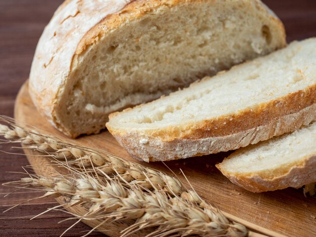 Foto close-up di pane di grano croccante fresco a fette giace su una tavola di legno sullo sfondo marrone di legno diverse orecchie di grano giacciono accanto al pane vista laterale