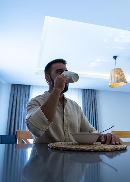 Closeup from below of young man with and shirt drinking from a white mug with a bowl of breakfast