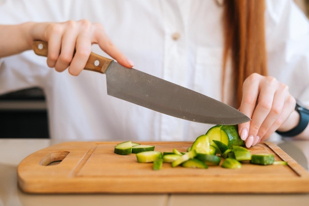 Photo closeup front view of unrecognizable woman cutting fresh cucumber cooking food salad sitting at wodden table in light kitchen room front view of female cooking vegetarian dieting salad at home