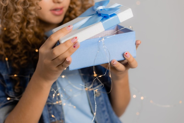 Primo piano di una confezione regalo tenuta nelle mani di una giovane donna bionda con i capelli ricci in abiti di jeans. la ragazza apre il regalo. fondo grigio solido. spazio per il testo. foto di alta qualità