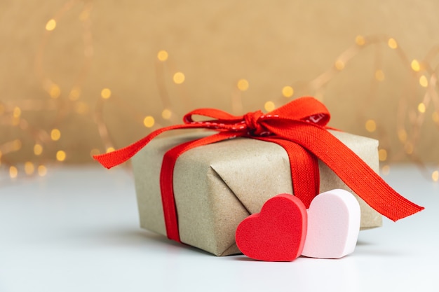 Closeup of a gift box with red ribbon and two hearts on blurred background