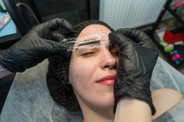 女の子の顔のクローズアップ 麻酔クリームが眉毛に塗布されます 美容師の手は透明なフィルムで眉毛を覆います