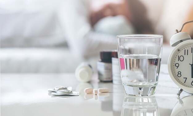 Closeup glass of drink water and pills on white table with  blurred background of man sleeping on sofa in living room.