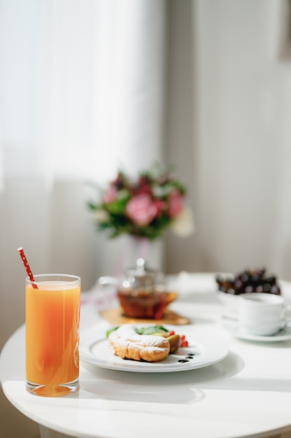 Photo closeup of a glass with juice and a drinking straw on a table with breakfast and a bouquet of