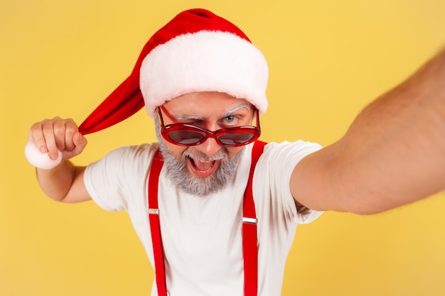 Closeup grappige oudere man in Kerstman hoed camera kijken en knipogen tonen tong plezier op wintervakantie Indoor studio opname geïsoleerd op gele achtergrond